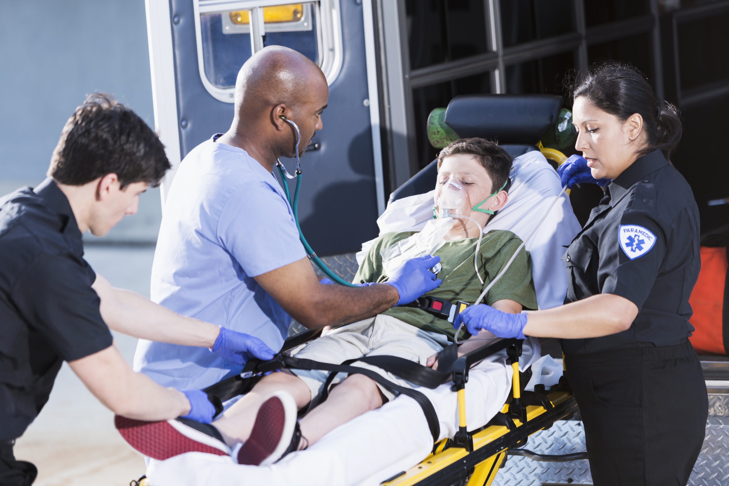 Multi-ethnic doctor and paramedics (30s, 40s) helping boy (12 years) lying on a stretcher outside an ambulance.